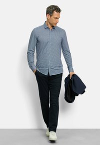 Hellblau kariertes Hemd mit Knöpfen, kombiniert mit dunklen Hosen, die mit einer marineblauen Jacke gehalten werden. Weiße Sneaker runden das Outfit ab.