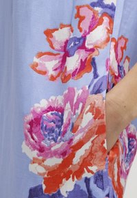 Robe florale ornée de grands motifs de fleurs roses et orange sur un tissu bleu clair. Inclut un détail de poche latérale.