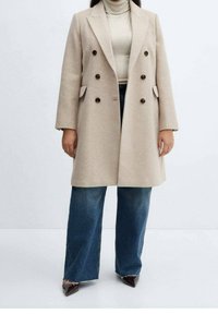 Beige uldblandet dobbeltradet frakke med spidse slag, mørke knapper og sidelommer, kombineret med en lysfarvet turtleneck og brede jeans.