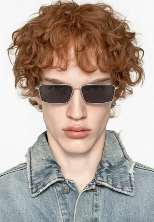 Jeune personne aux cheveux roux bouclés portant des lunettes de soleil rectangulaires foncées et une veste en jean bleu clair sur un fond uni.