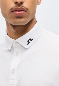 Άνδρας που φοράει λευκό πόλο με τρία κουμπιά και μαύρο γεωμετρικό λογότυπο στη δεξιά πλευρά του γιακά.