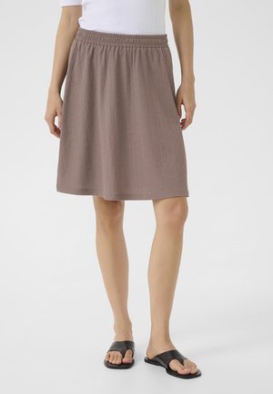 Vrouw met een bruine gestructureerde rok tot op de knie, wit topje en zwarte platte sandalen op een eenvoudige lichte achtergrond.