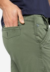 Green textured pants with a fitted design, featuring a side pocket and contrasting stitching. Paired with a navy shirt just above.