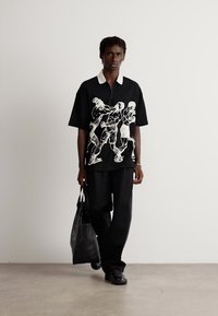 Man walking holding black tote bag, wearing black shirt with white illustrated figures, black pants, and black shoes against white background.