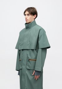 Zelená nepremokavá bunda s vysokým golierom, skrátenými rukávmi, dvoma vreckami so zipsom a oranžovými akcentmi na zipsoch. Hladká textúra, voľný strih.