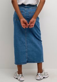 Jeansrock in einem mittleren Blauton, lang, mit geradem Schnitt, zwei Gesäßtaschen und einem Saum, der knapp über dem Knöchel endet. Getragen mit grauen Sneakern.