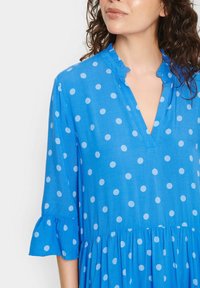 Blå klänning med vita prickar, med V-ringning och volangkrage, långa ärmar med utsvängda manschetter och en flödande kjol.