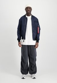 Marineblå bomberjakke med ribbede manchetter, hvid t-shirt under, løse mørkegrå bukser og hvide sneakers. Bærer en beige strikket beanie.