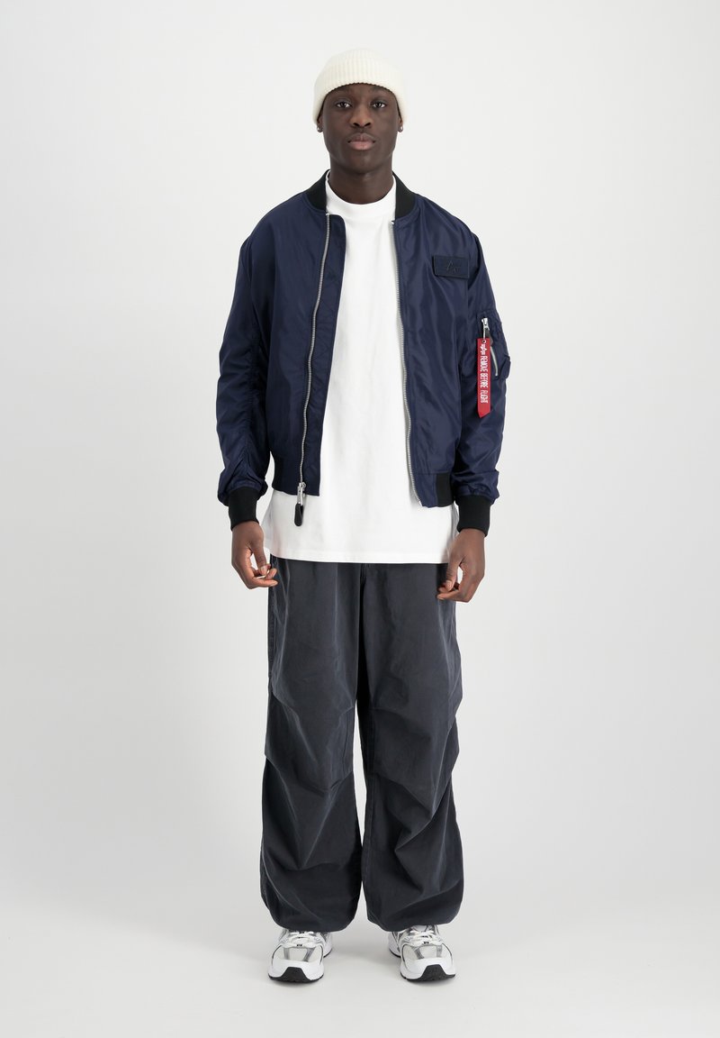 Marineblå bomberjakke med ribbede manchetter, hvid t-shirt under, løse mørkegrå bukser og hvide sneakers. Bærer en beige strikket beanie.