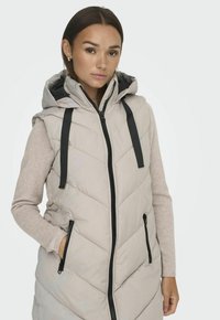Gilet matelassé beige clair avec capuche, présentant des coutures en chevrons, des zips noirs et des cordons de serrage, porté par-dessus un pull gris clair.