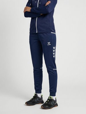 Osoba ubrana w granatowe spodnie i kurtkę dresowe hummel z białymi detalami w kształcie naszywek oraz czarne sneakersy z zielonymi akcentami, stojąca z założonymi ramionami.