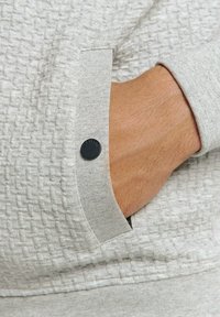 Lichtgrijze sweaterstof met textuur en een geweven patroon, featuring een manchetdetail en een zwarte ronde knoopaccent.