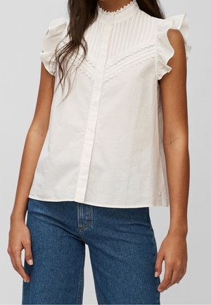 Kvinde iført en hvid ærmeløs bluse med flæser på skuldrene og blonde detaljer, kombineret med blå denimjeans.