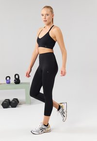 Black sports bra with a V-shaped neckline, paired with high-waisted black leggings. Includes grey and gold athletic shoes, kettlebells in background.