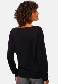Frau mit lockigem Haar, die einen schwarzen Pullover mit langen Ärmeln trägt und sich umgedreht hat, um den Rücken zu zeigen, kombiniert mit grauen Hosen.