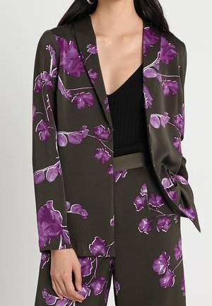 Vrouw draagt een donkergroene blazer en broek met paarse bloemenprint, gecombineerd met een zwart V-hals topje.