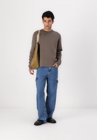 Smeđa sweatshirt, plave cargo traperice s bočnim džepovima i crne cipele. Model drži bež torbu, stojeći na bijeloj pozadini.