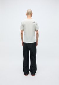 T-shirt gris clair à manches courtes avec un logo dans le dos, associé à un pantalon noir ample et des chaussures à accents rouges, vu de derrière.
