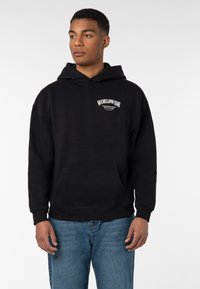 Junger Mann in schwarzem "WORLDWIDE" Hoodie und blauen Jeans, der mit entspannten Händen steht und leicht zur Seite schaut, vor weißem Hintergrund.