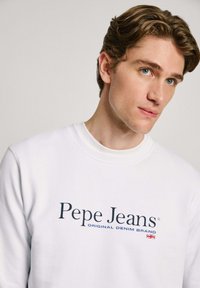 Sudadera blanca con logo y texto en azul marino "Pepe Jeans". Cuello redondo y puños de canalé, textura suave, diseño casual.