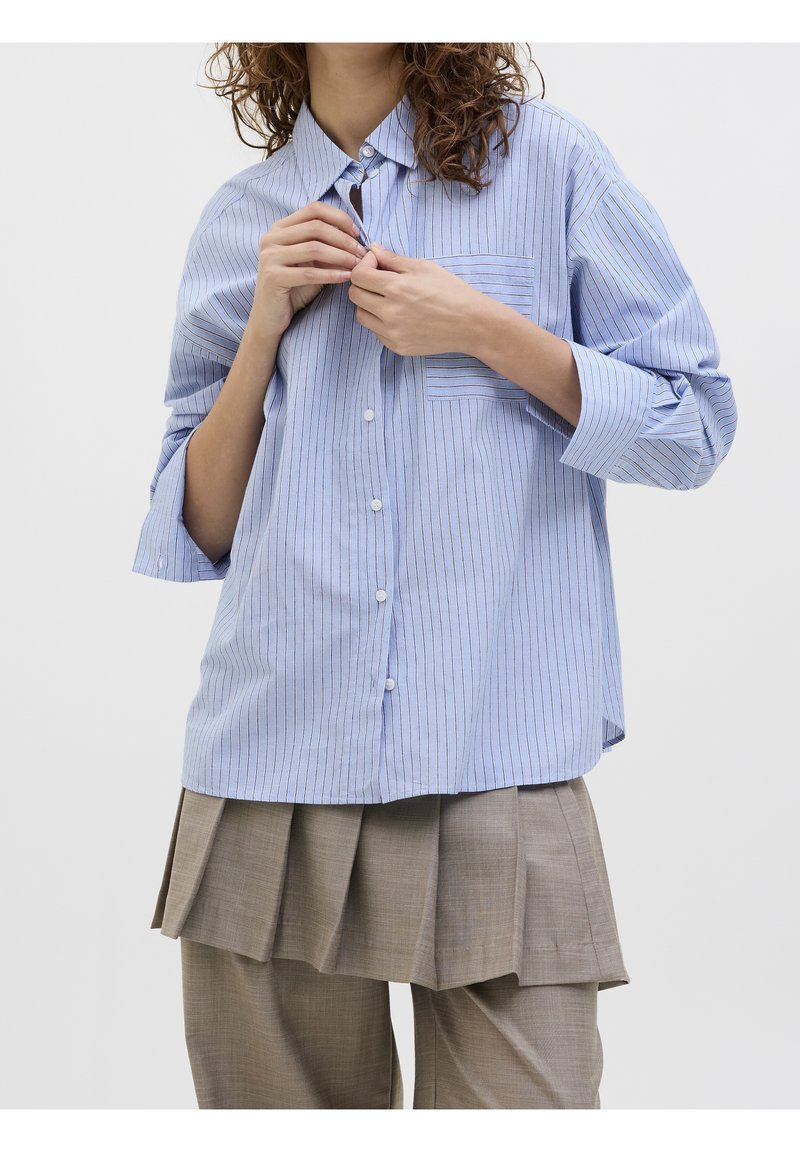 Femme aux cheveux bouclés en train de boutonner une chemise ample bleue à fines rayures, portant une jupe plissée beige par-dessus un pantalon ample.