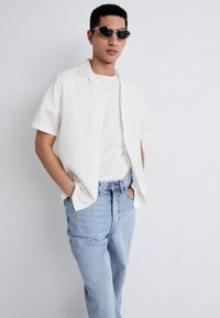 Chemise blanche à manches courtes avec un col camp, associée à un t-shirt blanc uni et un jean droit bleu clair, portée avec des lunettes de soleil noires.