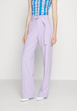 Frau trägt hoch taillierte lila weite Hosen mit Gürtel, blau-rosa kariertes ärmelloses Oberteil und weiße spitze Schuhe.