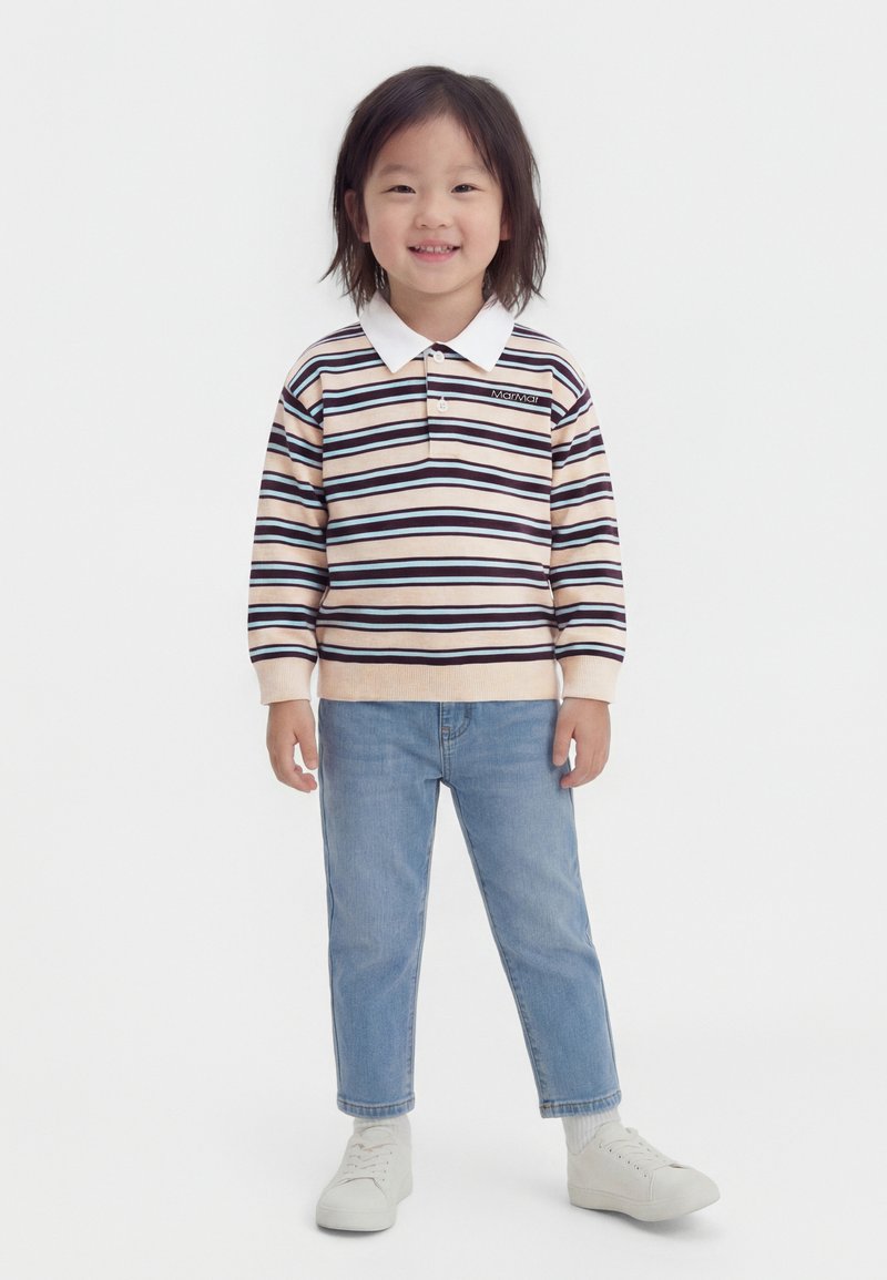 Χαμογελαστό παιδί που στέκεται, φοράει ριγέ μπλούζα πόλο με μακρύ μανίκι, ανοιχτό μπλε τζιν, λευκές κάλτσες και λευκά αθλητικά παπούτσια σε λευκό φόντο.