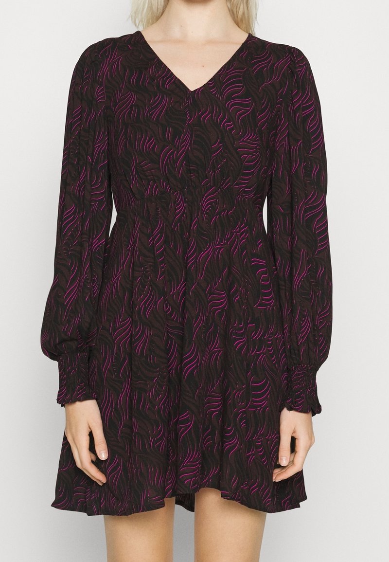 Femme portant une robe noire à manches longues avec des lignes ondulées abstraites violettes, une taille élastique, des poignets froncés et un col en V.