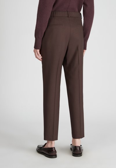 Persona che indossa pantaloni marroni su misura, mocassini marroni scuri e un maglione bordeaux, in piedi di fronte a uno sfondo chiaro e uniforme.