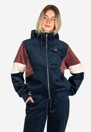 Young woman with blonde hair wearing a color-blocked zip-up hooded jacket and matching pants, hands in pockets, looking to the side.