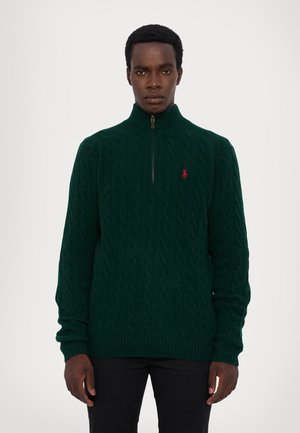 Grön stickad tröja med hög krage, featuring en fjärdedel dragkedja, kabelstickat mönster och en liten röd logotyp på bröstet.