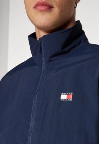 Granatowa kurtka wiatrówka z kołnierzem stojącym, pełnym zamkiem z przodu i małym haftowanym logo w kolorach czerwonym, białym i niebieskim. Tkanina o fakturze.