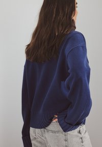 Navy blue knitted sweater with ribbed cuff and hem. The design features a relaxed fit, displayed with gray high-waisted jeans.