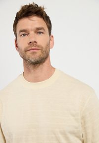 Sudadera de cuello redondo en beige claro con sutiles líneas de textura horizontal, hecha de un tejido suave. Presenta un cuello acanalado y mangas ajustadas.