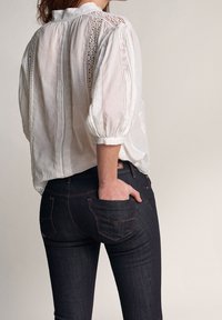 Vit, strukturerad blus med puffärmar och spetsdetalj, kombinerad med mörka denimjeans med bakficka och orange söm.