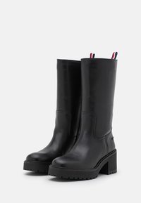 Bottes en cuir noir montantes avec un talon épais, bout rond, couture sur le côté et tirettes tricolores en haut. Semelle texturée.