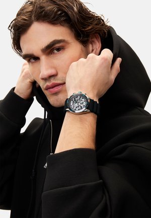 Montre chronographe numérique noire avec accents argentés et bracelet en caoutchouc noir, portée au poignet. Le modèle est vêtu d'un sweat à capuche noir et affiche un regard impassible.