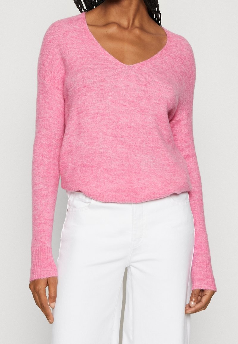 Femme portant un pull doux rose à col en V et un pantalon blanc, montrée des épaules aux hanches sur un fond uni.