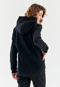 Černá mikina s kapucí, jemné textury, dlouhými rukávy, klokanní kapsou a žebrovanými manžetami, viděná zezadu, odpočívající na světlém pozadí.