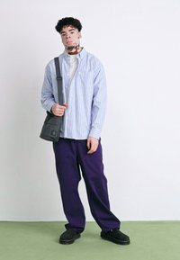 Stribet blå skjorte med knapper, grå taske, off-white skjorte under, lilla brede bukser og sorte sko, mod en hvid baggrund.