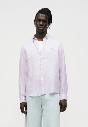 Młody mężczyzna o ciemnej karnacji i krótkich dredach, ubrany w jasnofioletową koszulę zapinaną na guziki na białej koszulce i jasnoniebieskich jeansach, stojący na białym tle.