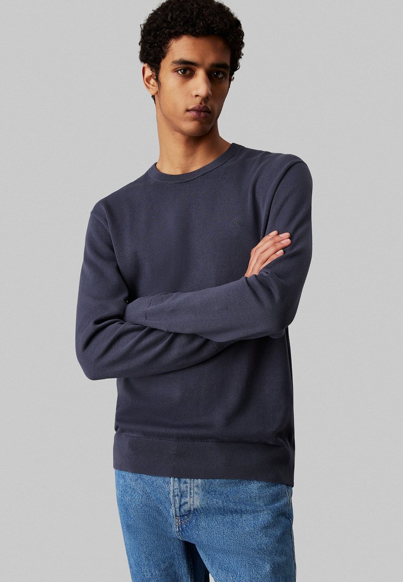 Maglione a maglia blu navy con colletto a girocollo a coste, maniche lunghe e orlo elastico. Presenta un sottile dettaglio del logo sul petto. Indossato con jeans blu.