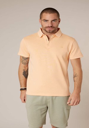 Homme aux cheveux courts et tatouages, portant un polo pêche et un short vert clair, debout devant un fond beige uni.