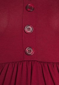 Vestido vermelho com um corpete justo e três botões redondos e transparentes. A saia possui uma textura franzida, adicionando volume e detalhe.