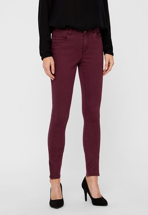 Kobieta w bordowych obcisłych dżinsach z kieszeniami z przodu i z tyłu, w połączeniu z czarną długą bluzką i czarnymi szpilkami.