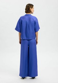 Mėlyna laisva marškinė ir plačios kelnės, pagamintos iš lygaus audinio. Marškiniai turi apykaklę ir nugaroje įliekimą, su trumpomis rankovėmis.