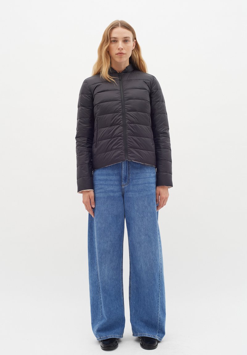 Schwarze Daunenjacke mit hohem Kragen, horizontal gestepptem Design, Frontreißverschluss, kombiniert mit weit geschnittenen blauen Jeans.