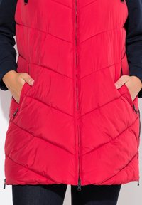 Rote gepolsterte Weste mit Zickzackmuster, Frontreißverschluss und Seitentaschen, mit einer glatten Textur und abgerundetem Saum.