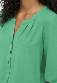 Blusa verde con una texture liscia, caratterizzata da un collo rotondo, chiusura a bottoni e sottili pince davanti.